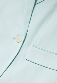 Veste bleu clair avec un seul bouton beige et une poche passepoilée sur le côté droit à l'avant.