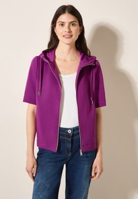 Lila zip-up hoodie med korta ärmar, med huva och dragskor. Bärs över en vit topp, i kombination med blå denimbyxor.