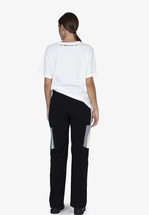 T-shirt bianco in cotone con etichetta stampata sul retro, abbinato a pantaloni neri con pannelli grigi ai lati.