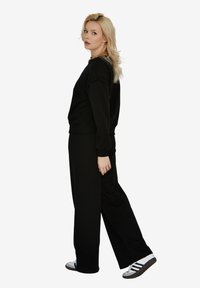 Czarny sweatshirt i szerokie spodnie wykonane z miękkiego materiału. Modelka nosi czarno-białe sneakersy oraz złoty naszyjnik.
