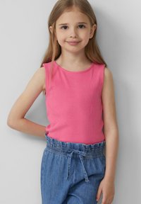 Jong meisje met lang blond haar, gekleed in een mouwloos roze topje en blauwe hoge taille spijkerbroek, lachend met één hand achter haar rug.