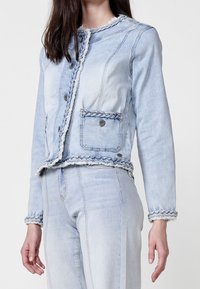 Light blue denim jacket with braided accents, frayed edges, button closure, and two front pockets. Long sleeves and tailored fit.