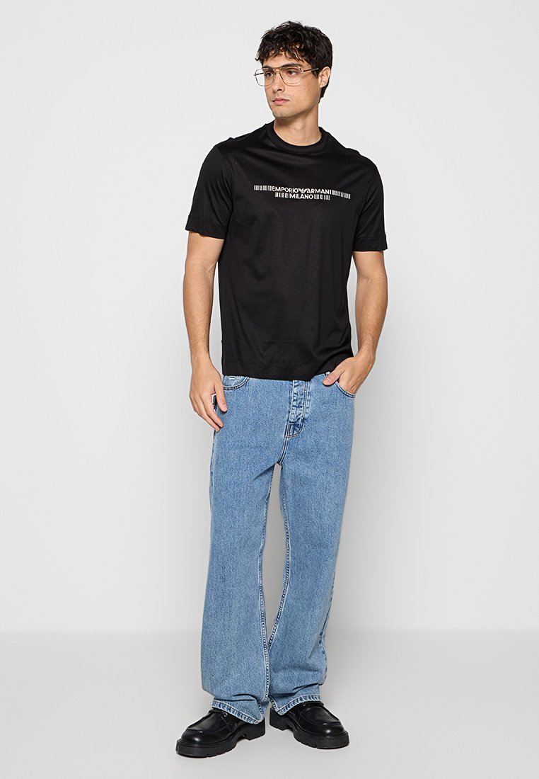 Hombre con camiseta negra Emporio Armani Milano, vaqueros azul claro, zapatos negros y gafas, de pie con una mano en el bolsillo.