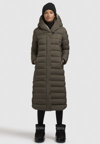 Garš olive pelēks pūkains mētelis ar kapuci, pogām, sānu kabatām un horizontālu šūšanas rakstu, apvienots ar melnajām zābakiem.