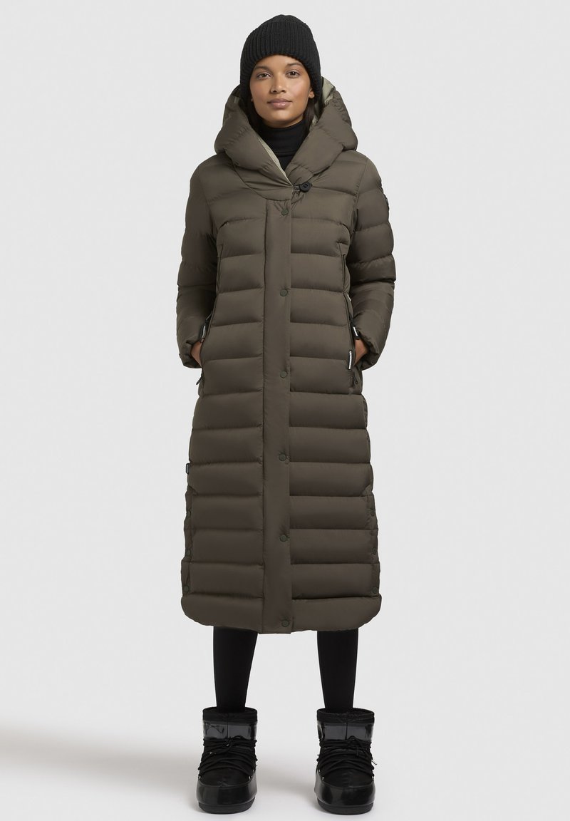 Garš olive pelēks pūkains mētelis ar kapuci, pogām, sānu kabatām un horizontālu šūšanas rakstu, apvienots ar melnajām zābakiem.