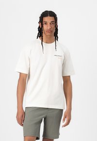 Cremefarvet bomulds-T-shirt med korte ærmer, forsynet med McLaren-logo på brystet. Kombineret med strukturere olivengrønne shorts.
