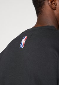 Gros plan d'une personne portant un t-shirt noir avec le logo NBA imprimé sur le haut du dos près de l'encolure.