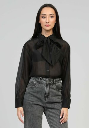 Blouse noire transparente avec un grand nœud au col, devant à boutons, et manches longues. Associée à un jean en denim gris taille haute.