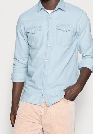 Homme portant une chemise en denim bleu clair boutonnée avec manches retroussées et un pantalon en velours côtelé rose pâle, main dans la poche.