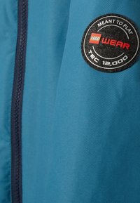Blaue Jacke aus strukturiertem Stoff, mit sichtbarem Reißverschluss und einem runden schwarz-roten Logo-Patch auf dem Ärmel, auf dem "LEGO WEAR TEC 12.000" steht.