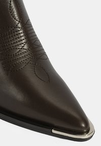 Botte en cuir marron à chevilles avec un bout pointu, décorée de coutures et d'un accent en capuchon argenté sur une semelle extérieure noire.