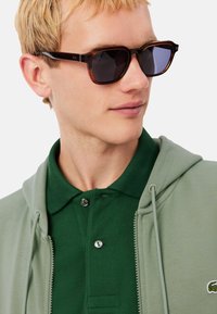 Gafas de sol con montura de carey, lentes oscuras, forma rectangular y detalle sutil de logo. Usadas con un polo verde y una sudadera con capucha verde clara.