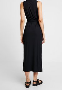 Czarna, bezrękawnikowa sukienka midi z dopasowaną talią i bocznymi rozcięciami. Gładki, elastyczny materiał. Noszona z czarnymi sandałami, widoczne tatuaże na kostkach.
