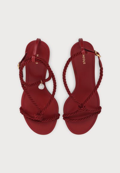 Paire de sandales rouges avec des brides en corde torsadée, boucle dorée et un petit charm blanc décoratif sur une bride, sur un fond clair.
