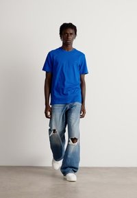 Camisa de algodón azul con mangas cortas, combinada con vaqueros desgastados de color azul claro y zapatillas blancas, sobre un fondo blanco liso.