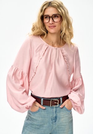 Blonde vrouw met grote schildpadkleurige bril, lichtroze blouse met ruches en pofmouwen, ingestopt in lichtblauwe jeans met een bruine riem.