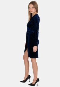 Abito in velluto blu navy con maniche lunghe e design avvolgente. Presenta uno spacco alto fino alla coscia e una silhouette aderente, abbinato a tacchi alti neri.