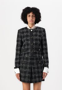 Mustavalkoinen ruudullinen bleiseri, jossa on jämäkkä design, kultasävyiset yksityiskohdat ja erottuvat valkoiset hihansuut. Karkea kangas, jossa on taskut.