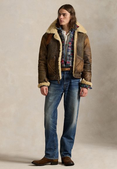 Giovane persona che indossa una giacca di pelle con shearling, camicia a quadri, jeans blu e stivali marroni, in piedi contro uno sfondo neutro.