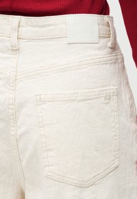 Κοντινό πλάνο της πίσω τσέπης και της μέσης μιας παντελόνας μπεζ υφής, που φοριέται με κόκκινο ριμπ πουλόβερ μέσα.