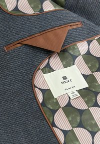 Hallvärvi tekstureeritud kangas pruunide servadega, mustrilise voodriga, millel on rohelised ja roosad ringid. Silt ütleb "NEXT, Slim Fit, 40R, Made in Vietnam."