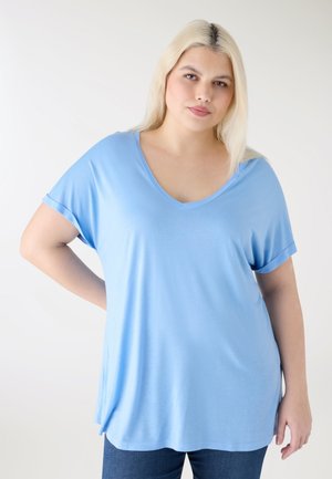 Blondynka w jasnoniebieskiej bluzce z krótkim rękawem i dekoltem w serek oraz ciemnych dżinsach, stojąca z jedną ręką na biodrze na białym tle.