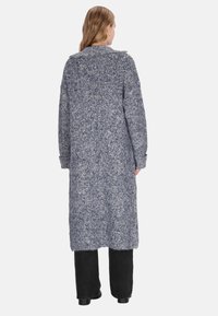 Donna che indossa un lungo cardigan lavorato a maglia blu-grigio, pantaloni neri e scarpe nere, in piedi di spalle su uno sfondo bianco.