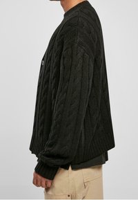 Svart stickad cardigan med ett kabelstickat mönster, ribbade muddarna och rund halsringning. Stängs med en enkel knappdetalj.