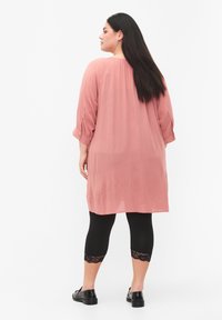 Vaaleanpunainen, löysä tunika, jossa on kolme neljännesosaa hihat, yhdistettynä mustiin legginseihin, joissa on pitsiä hihansuissa, ja mustiin kenkiin.