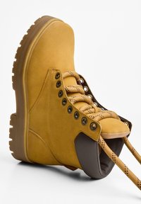 Gelbe Wildleder-Stiefelette mit gepolstertem Schaft, braunen Akzenten, metallischen Ösen und strukturierten Schnürsenkeln. Gummisohle mit tiefem Profil.