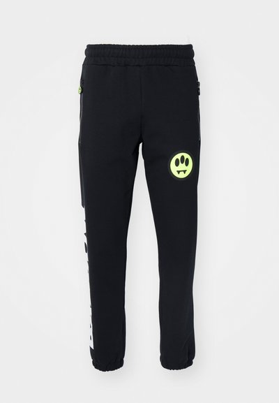 Black jogger pants made of soft fabric, featuring a neon green smiley face and white graphic print on the side, with elastic cuffs and waistband.