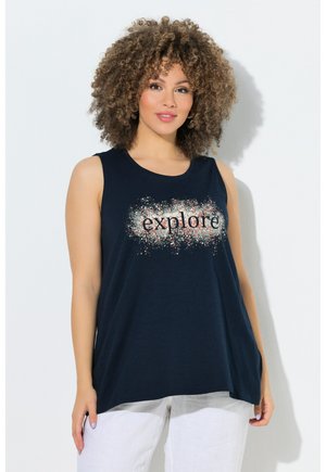 Frau mit lockigem Haar, die ein ärmelloses marineblaues Top mit dem Schriftzug „explore“ und weiße Hosen trägt, steht vor einem schlichten hellen Hintergrund.
