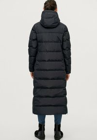 Lange zwarte pufferjas met capuchon, gewatteerd patroon en een getailleerde pasvorm. Heeft zijritsen en ribgebreide manchetten. Gelaagd materiaal.