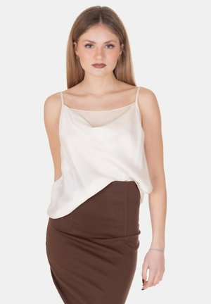 Giovane donna con capelli lunghi che indossa un top bianco senza maniche e una gonna marrone aderente, in piedi davanti a uno sfondo bianco uniforme.