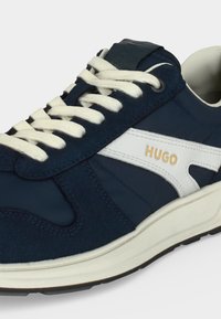 Baskets bleu marine avec lacets blancs, bande latérale blanche, semelle beige et logo « HUGO » doré sur le côté.