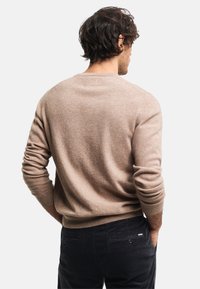 Hellbeige Pullover mit Rundhalsausschnitt aus weichem Material, mit gerippten Bündchen und Saum, getragen über dunklen Hosen. Die Ansicht zeigt das Rückendesign und die Textur.