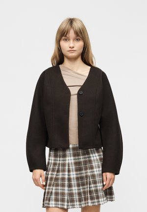 Νεαρή γυναίκα που φοράει μαύρο σακάκι με κουμπιά πάνω από μπεζ τοπ και πλισέ καρό φούστα σε καφέ και άσπρο, στέκεται μπροστά σε απλό φόντο.