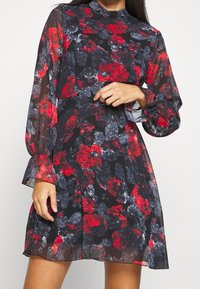 Un vestido floral con un fondo negro, patrones rojos y grises, mangas largas transparentes, un escote alto y un dobladillo acampanado.