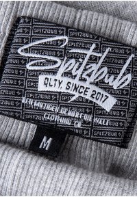 Grahamstoffetikett mit der Marke "Spitzbub" in weißer Stickerei. Beinhaltet den Text "Qualität seit 2017" und die Größenangabe "M" auf einem gerippten Hintergrund.