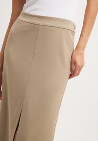 Beige Midi-Rock mit glatter Textur, seitlichem Schlitz und schmalem Bund, dargestellt mit einer Hand, die den Stoff hält.