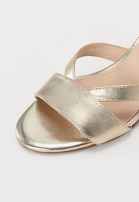 Gullgyll metallisk åpen tå sandal med bred kryssremdesign, glatt tekstur og tan fotseng, med lav flat hæl.