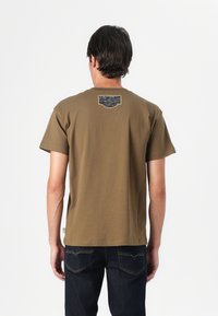 Brun bomull t-skjorte med rund hals og korte ermer. Har en lapp med et blått og svart design på ryggen. Brukes med mørke jeans.