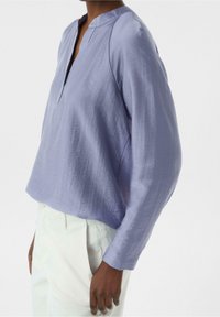 Chemise à manches longues lavande avec une coupe décontractée, présentant une texture subtile et un col en V. Associée à un pantalon de couleur claire.