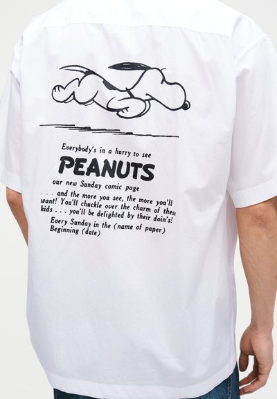 Καρτούν με τον Σνούπι να τρέχει πάνω από κείμενο που προωθεί το κυριακάτικο κόμικ Peanuts στην πίσω πλευρά ενός λευκού κοντομάνικου που φοριέται από ένα άτομο.