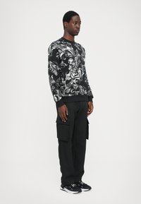 Maglione nero con motivo ondulato argentato, scollatura rotonda, polsini a coste. Abbinato a pantaloni cargo neri e scarpe sportive nere.