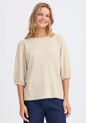 Beige karierte Bluse mit Dreiviertelärmeln, Rundhalsausschnitt und weicher Textur; kombiniert mit dunkelblauen Jeans.