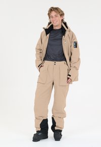 Beige vedenpitävä hiihtotakki mustalla sisäpuolella ja logolla, yhdistettynä yhteensopiviin beigen värisiin housuihin, joissa on taskuja ja mustia yksityiskohtia.