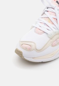 Baskets blanches de face avec des accents beige et rose pastel, lacets blancs et semelle texturée sur fond blanc.