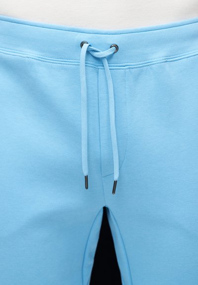 Φωτεινό γαλάζιο παντελόνι φόρμας με κορδόνι στη μέση και μεταλλικά ροκανίδια, εμφανιζόμενο από τη μέση έως το πάνω μέρος του μηρού από μπροστά.