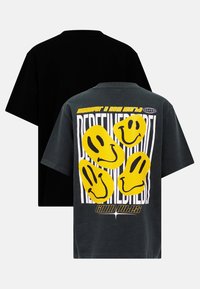 Svart och grå oversized T-shirt i bomull med gula leende ansiktstryck och textgrafik på baksidan, korta ärmar och en klassisk rund halsringning.
